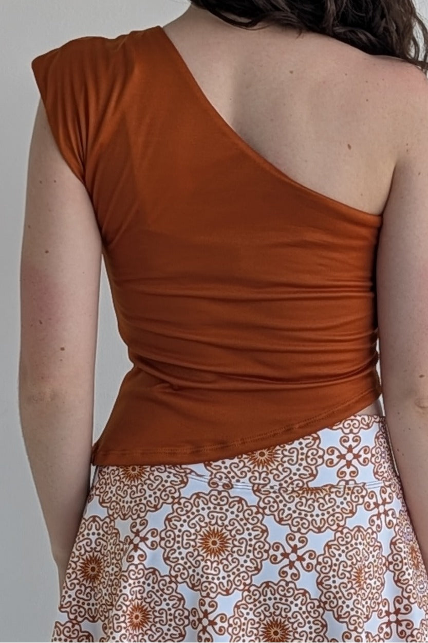 Person wearing a burnt orante one-shoulder top with a patterned skirt.