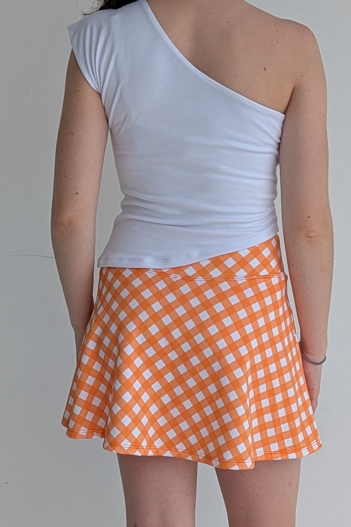 Person wearing a white one-shoulder top and orange checkered skirt on a plain background