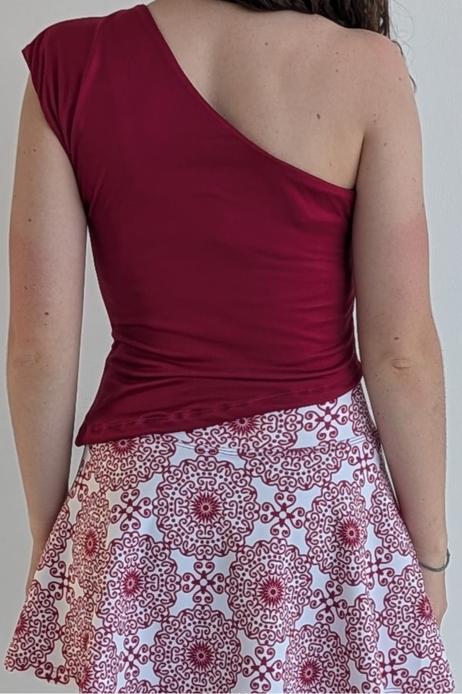 Person wearing a red one-shoulder top and patterned skirt against a plain background